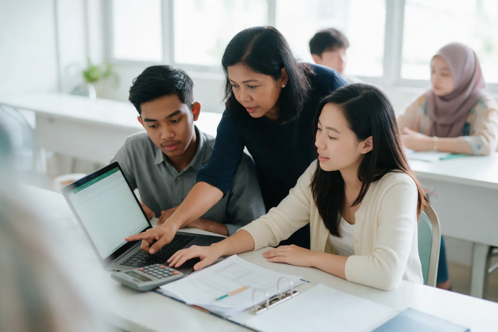 A lecturer leaning in between two students to point at a laptop in a bright accounting classroom