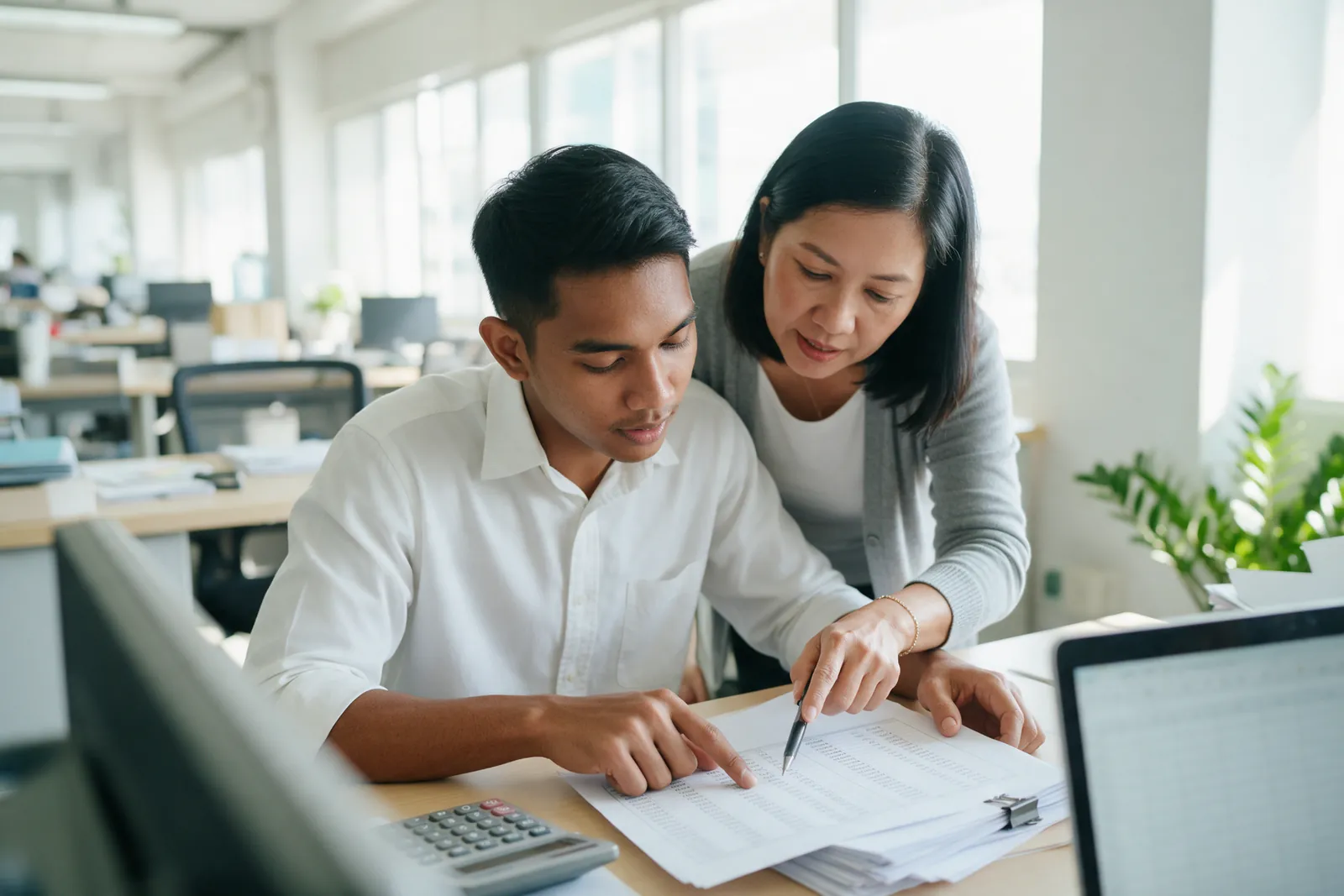 A trainee at a finance desk reviewing a printed sheet with a senior colleague pointing with a pen