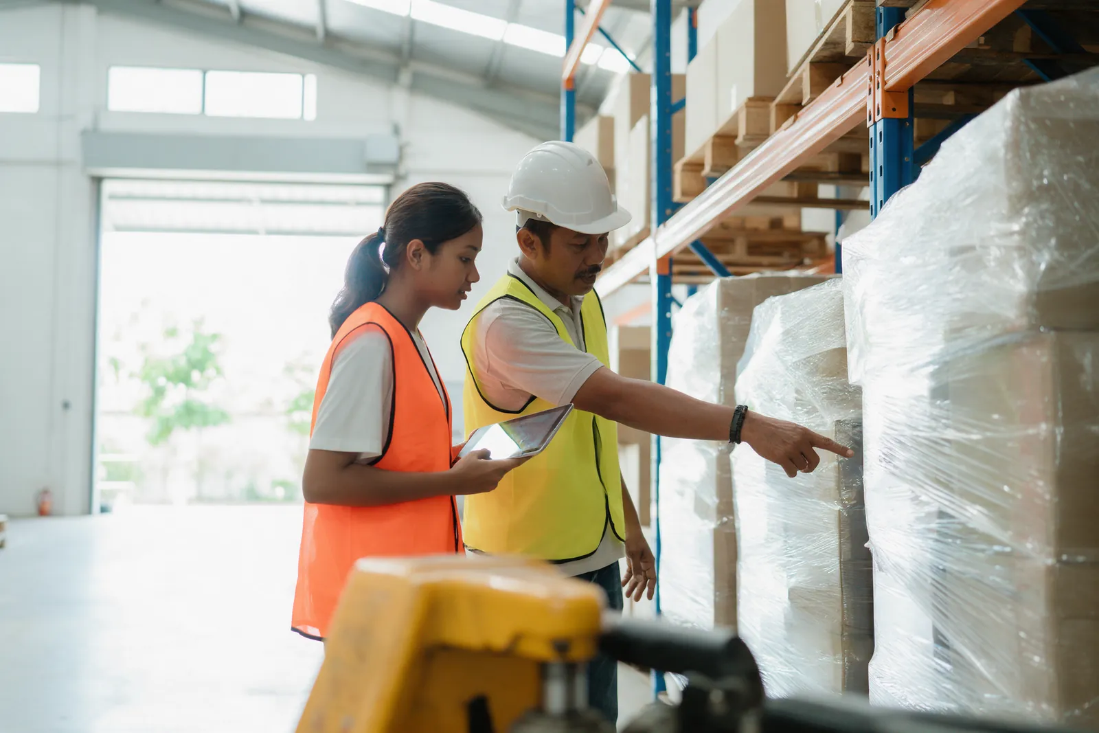 A trainee in a hi-vis vest holding a tablet while a supervisor points down a warehouse aisle